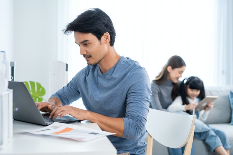 Asian business man father sit on table work at home with family behind in living room at house.
